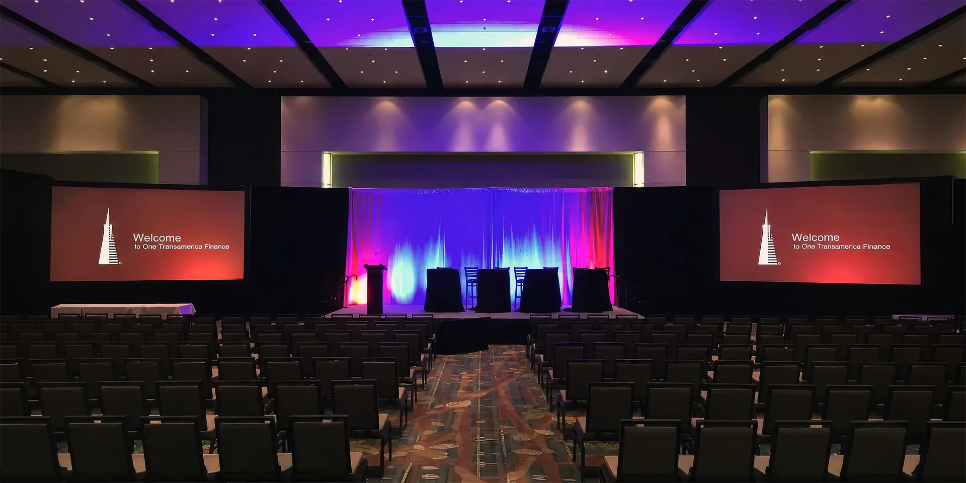 Hotel ballroom event setup with two large projection screens, stage lighting and audience seating for a corporate presentation.