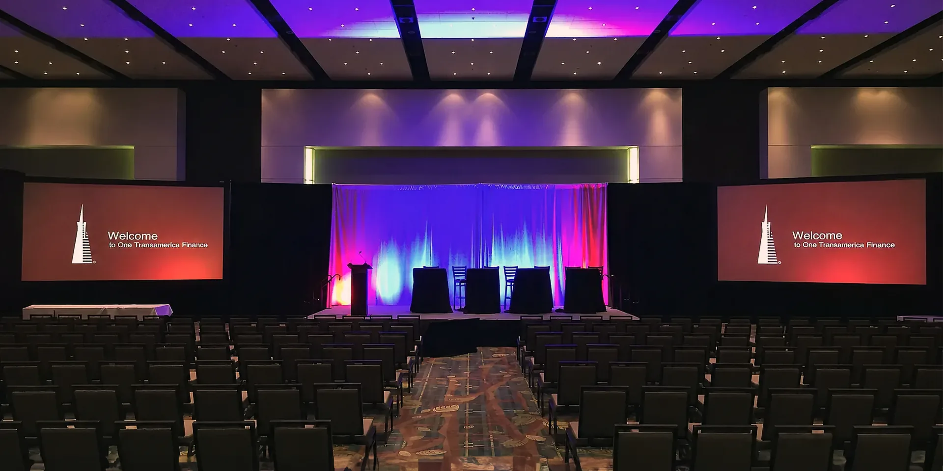Hotel ballroom AV setup with dual projection screens, a presentation stage and conference seating.