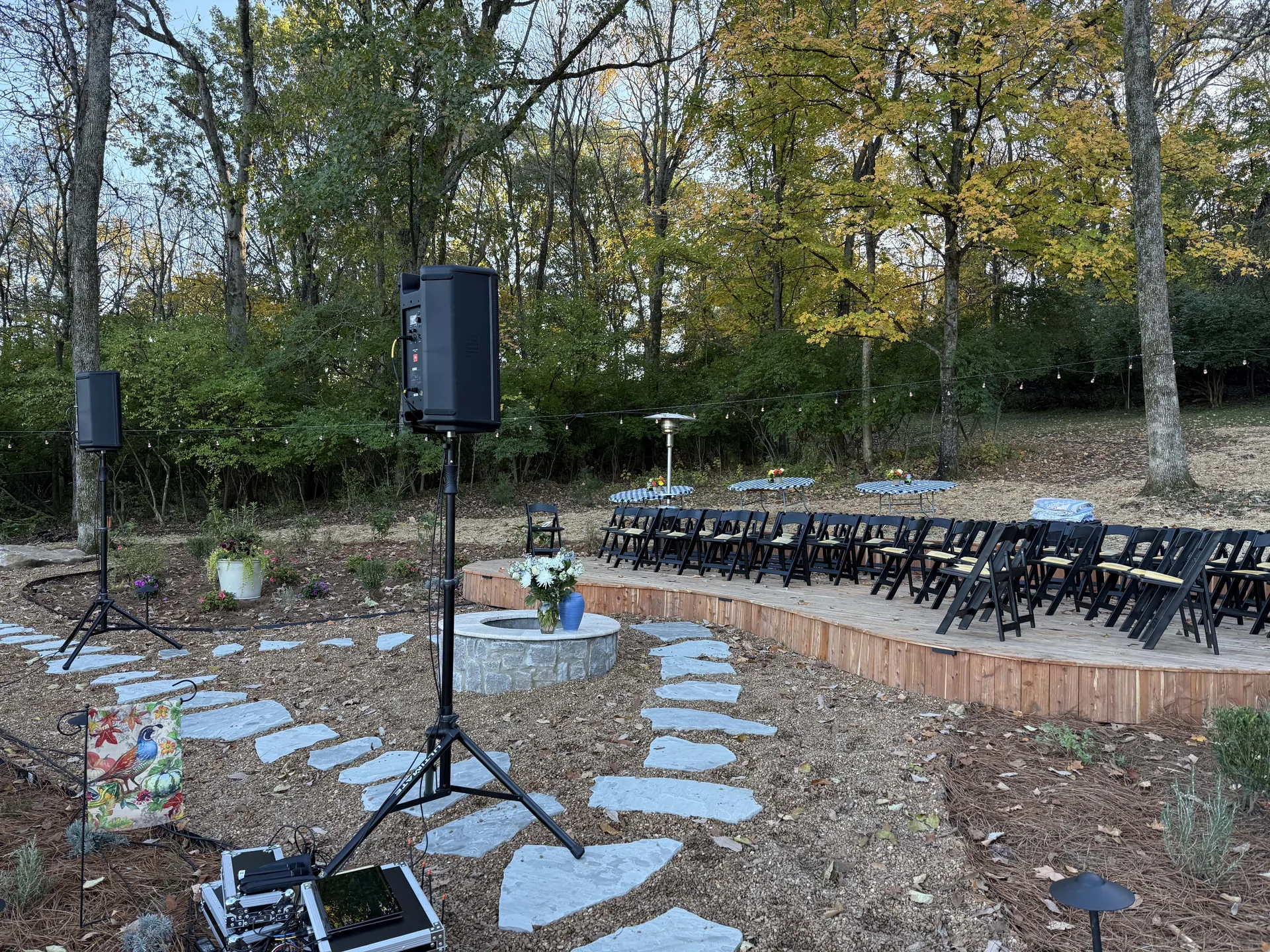 Outdoor celebration of life audio setup with black speakers on stands and seating in a wooded area