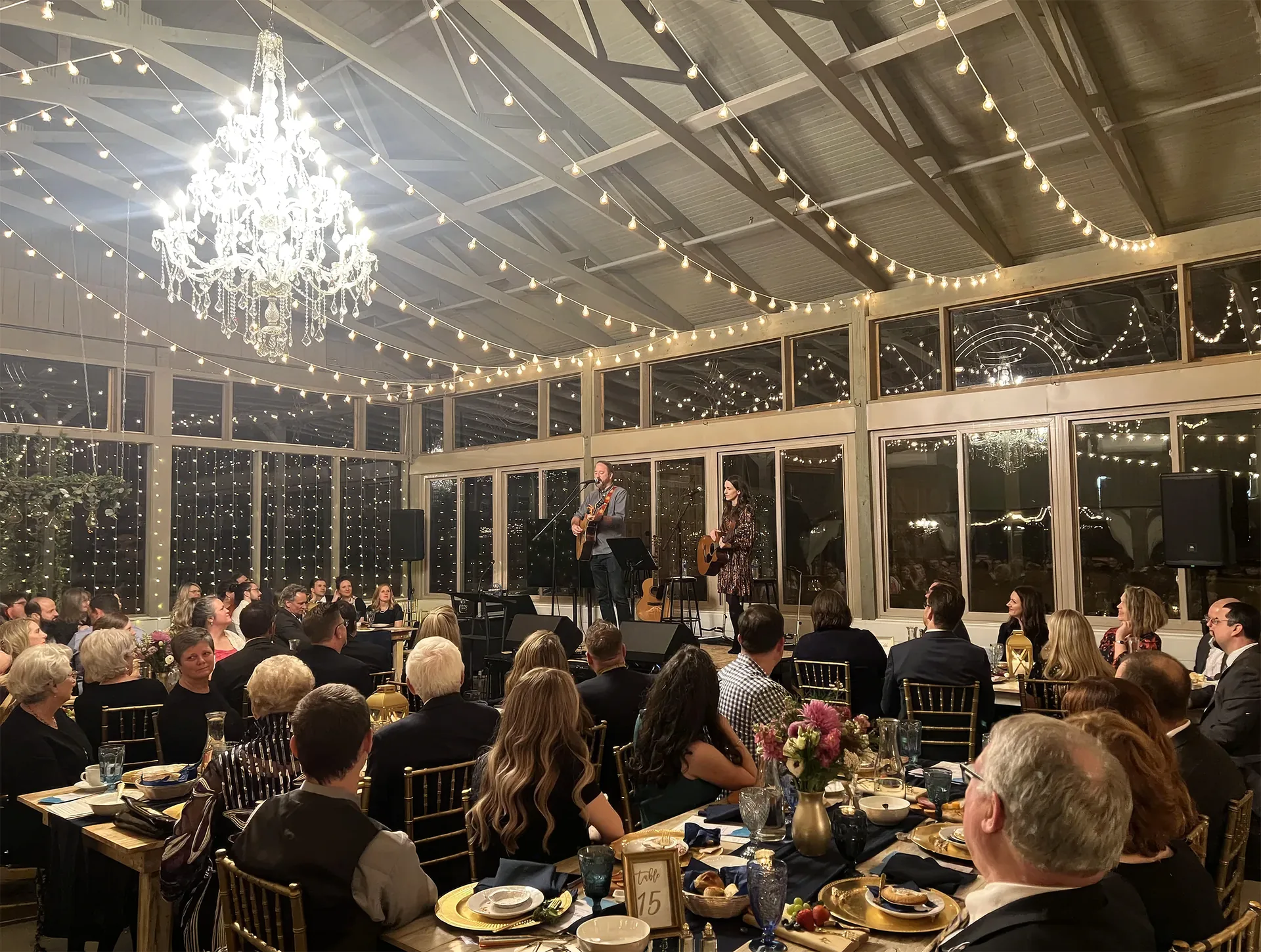 Elegant evening gala in a glass-enclosed space with guests enjoying live music under string lights