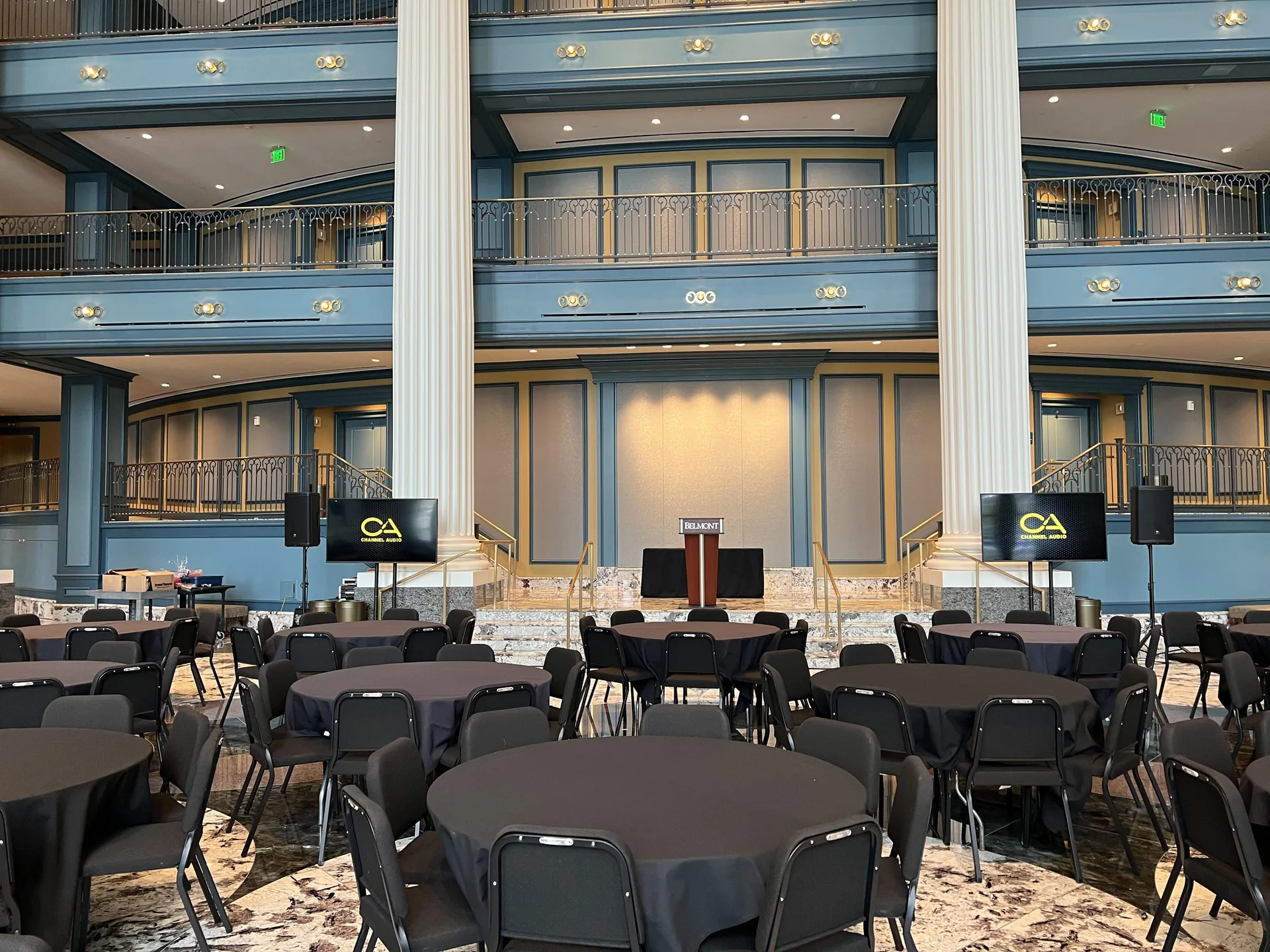 Grand hall event setup with two TV displays, PA speakers, podium and round-table seating