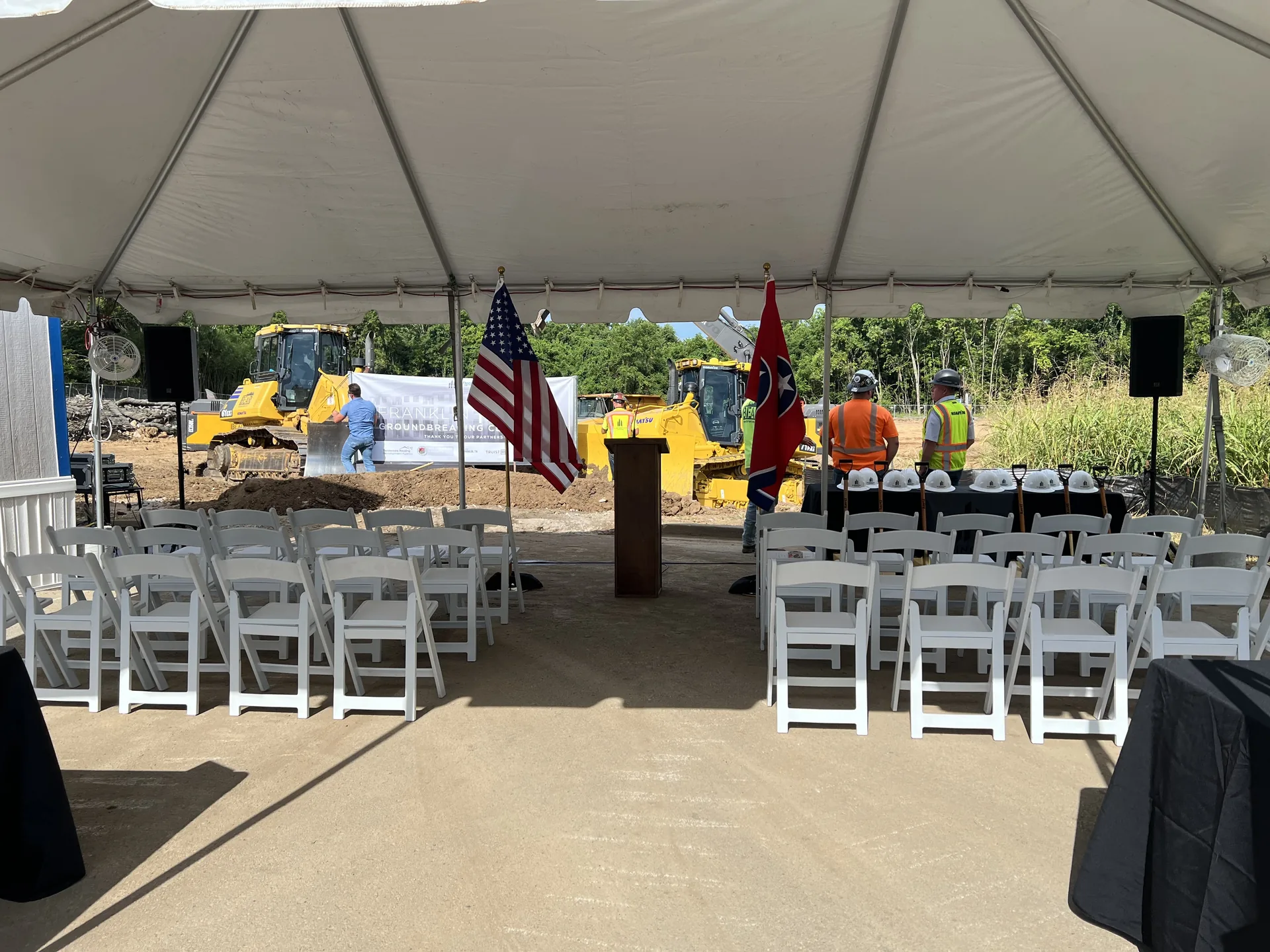 Groundbreaking ceremony under a tent with podium, flags, white seating and PA speakers