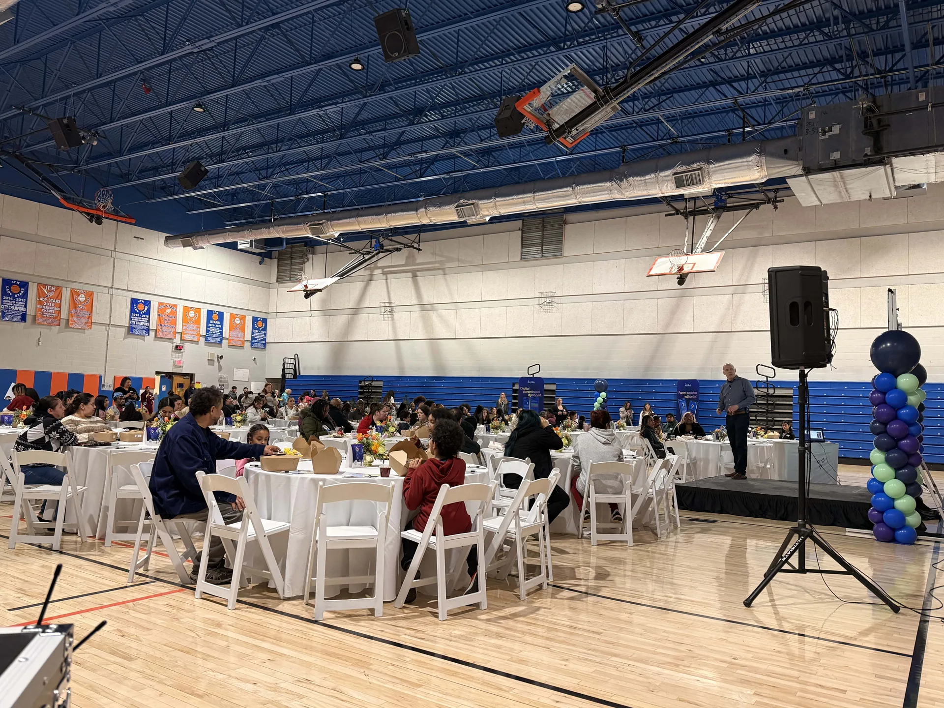 Gymnasium banquet with a PA speaker on a stand serving guests seated at round tables