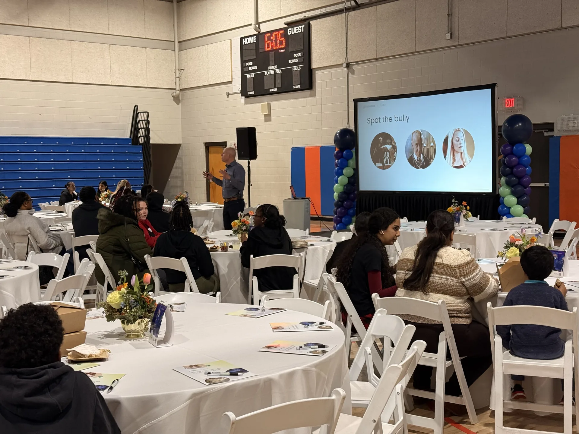 Presenter speaking in a gymnasium with a projection screen, PA system and balloon columns