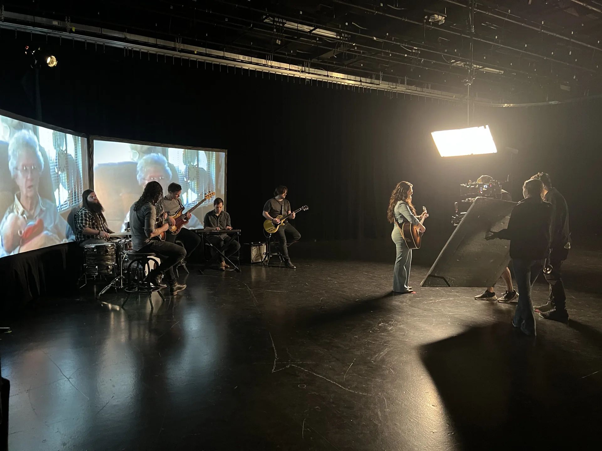 Live band recording session with multiple musicians and large screen displaying footage in background