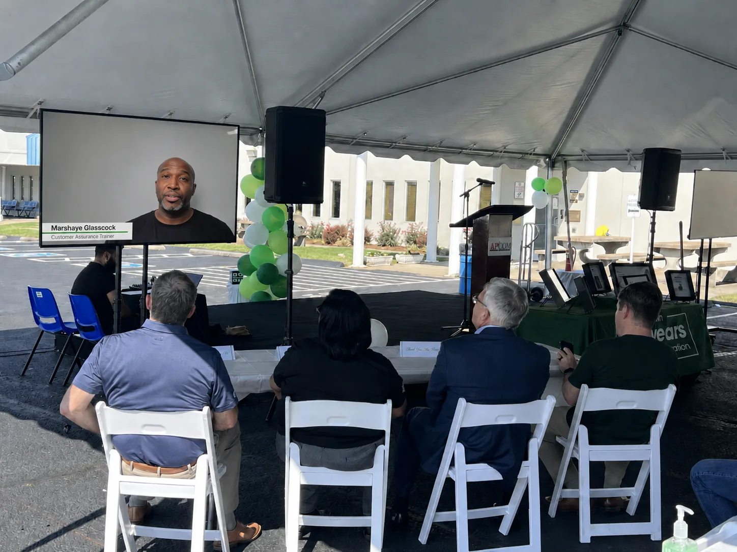 Outdoor corporate event setup with a large TV and speakers under a tent, audience seated in white chairs
