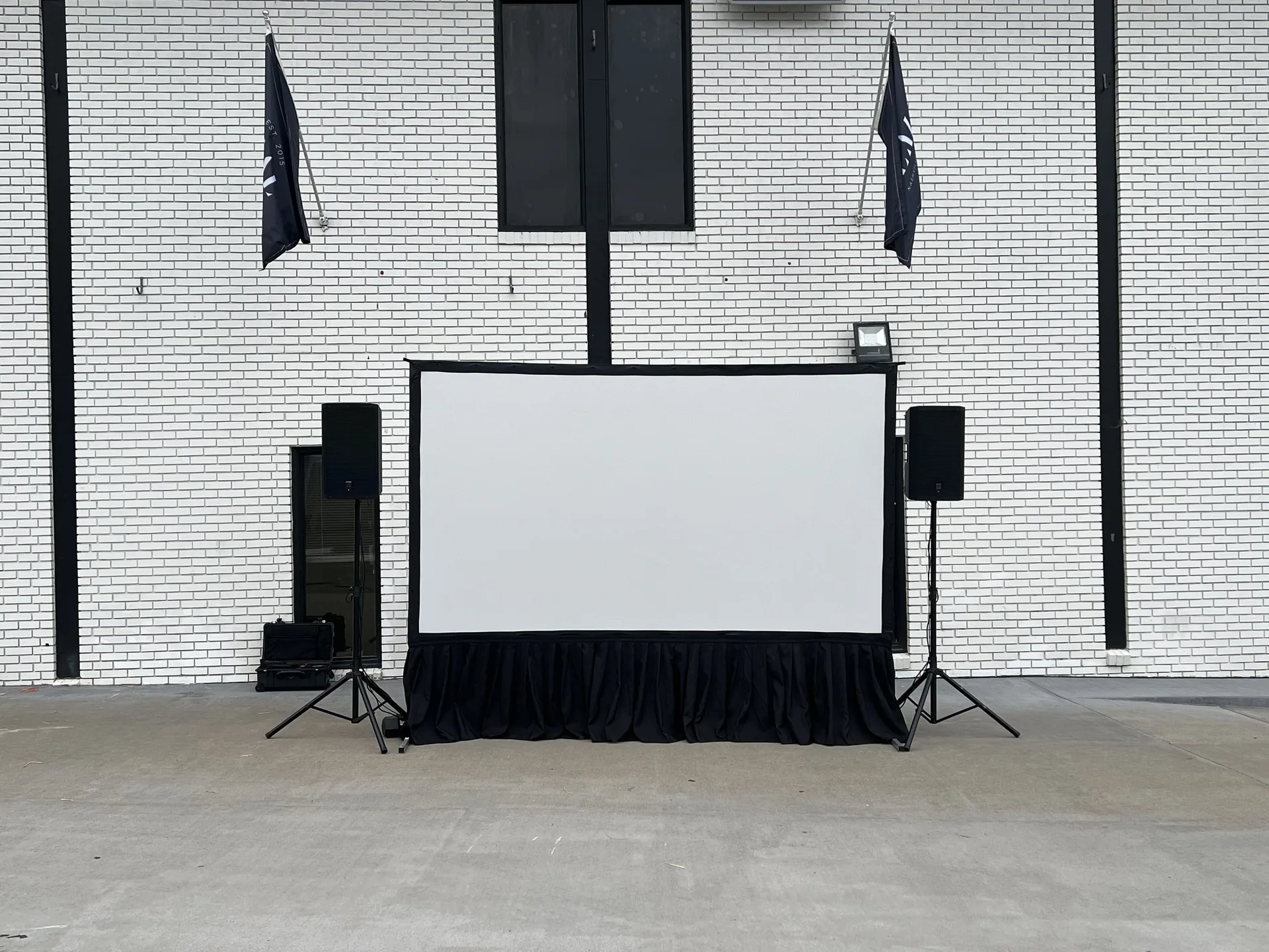 Outdoor movie screening setup with a large screen and speakers in front of a white brick building