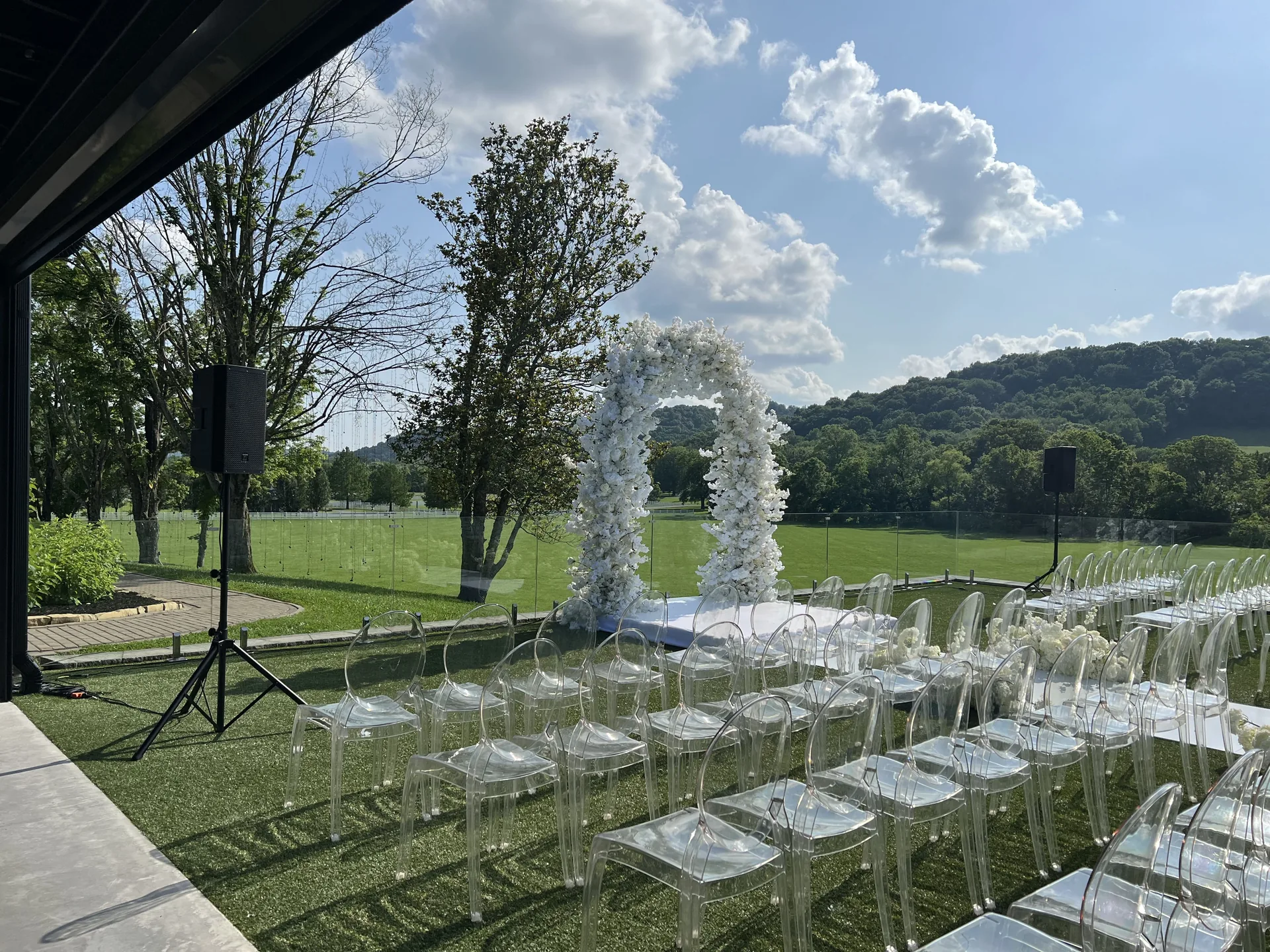 Outdoor wedding ceremony with PA speaker, clear chairs and white floral arch overlooking a green landscape