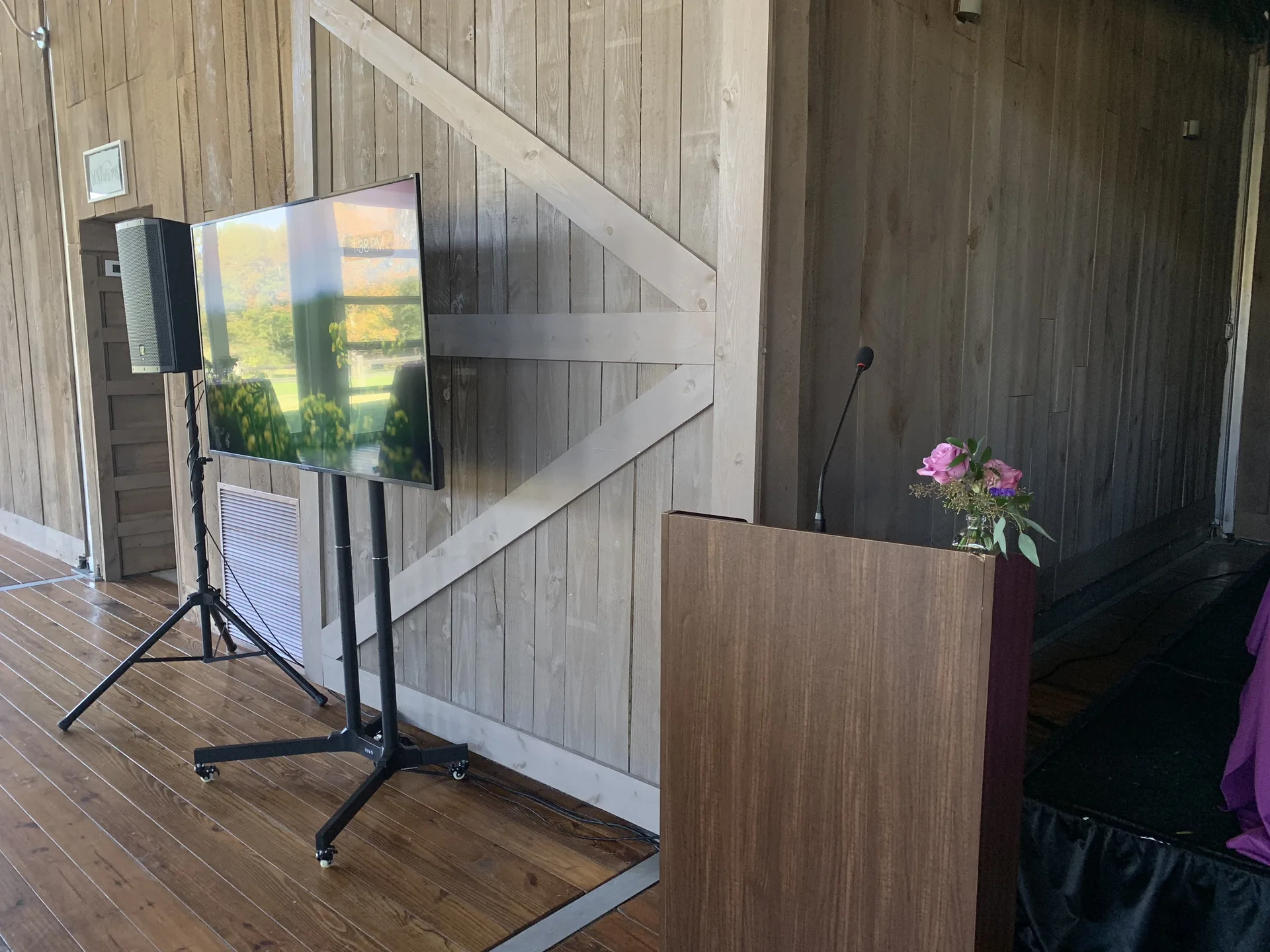Seminar setup with a TV display on a floor stand, podium and PA audio equipment beside a rustic wood wall